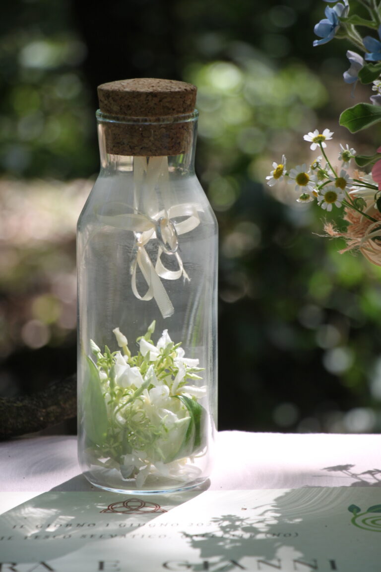 Un'elegante bottiglia di vetro con tappo in sughero, sospende delicatamente due anelli nuziali tramite nastri in raso avorio, con fiori freschi di gelsomino alla base. Un tocco naturale ed elegante per un matrimonio eco-friendly.