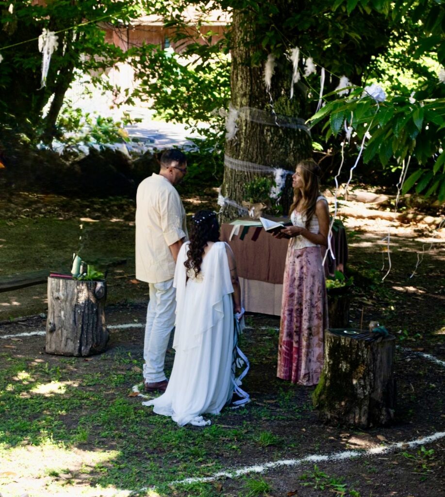 Cerimonia di matrimonio nel bosco sopra Genova, in Liguria, durante una giornata di settembre. La coppia è protagonista di un rito ispirato al matrimonio celtico, immerso nella natura tra alberi, foglie e muschio. La sposa indossa un abito bianco elegante e fluido, lo sposo un completo bianco raffinato. L’atmosfera è intima e naturale, con amici e parenti riuniti attorno alla coppia in un contesto boschivo luminoso e silenzioso. Il rito si svolge all’aperto, in un ambiente suggestivo che valorizza la natura e la semplicità della cerimonia.